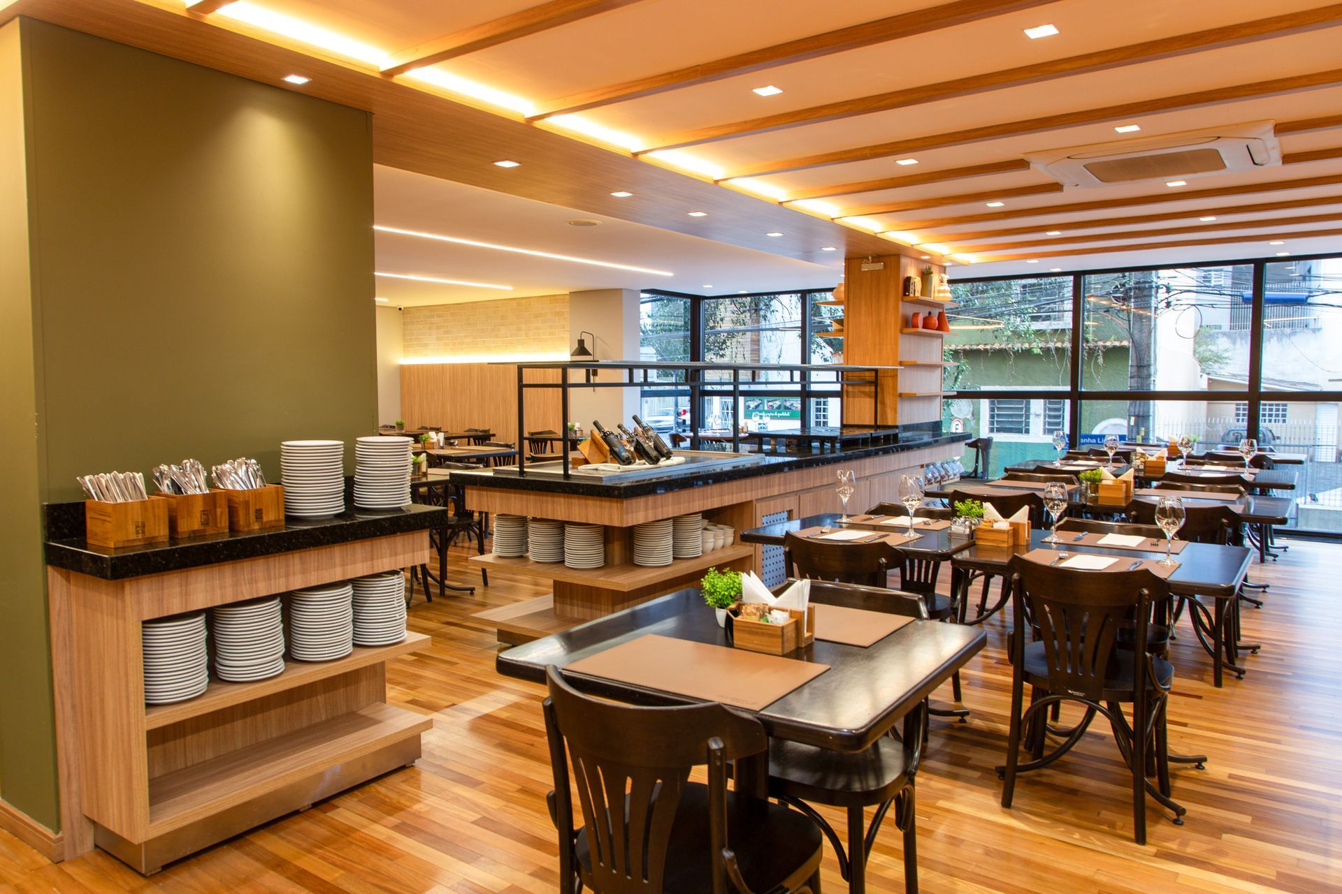 Área de jantar de restaurante iluminada com mesas e cadeiras de madeira, e um buffet com pratos e tigelas empilhados.