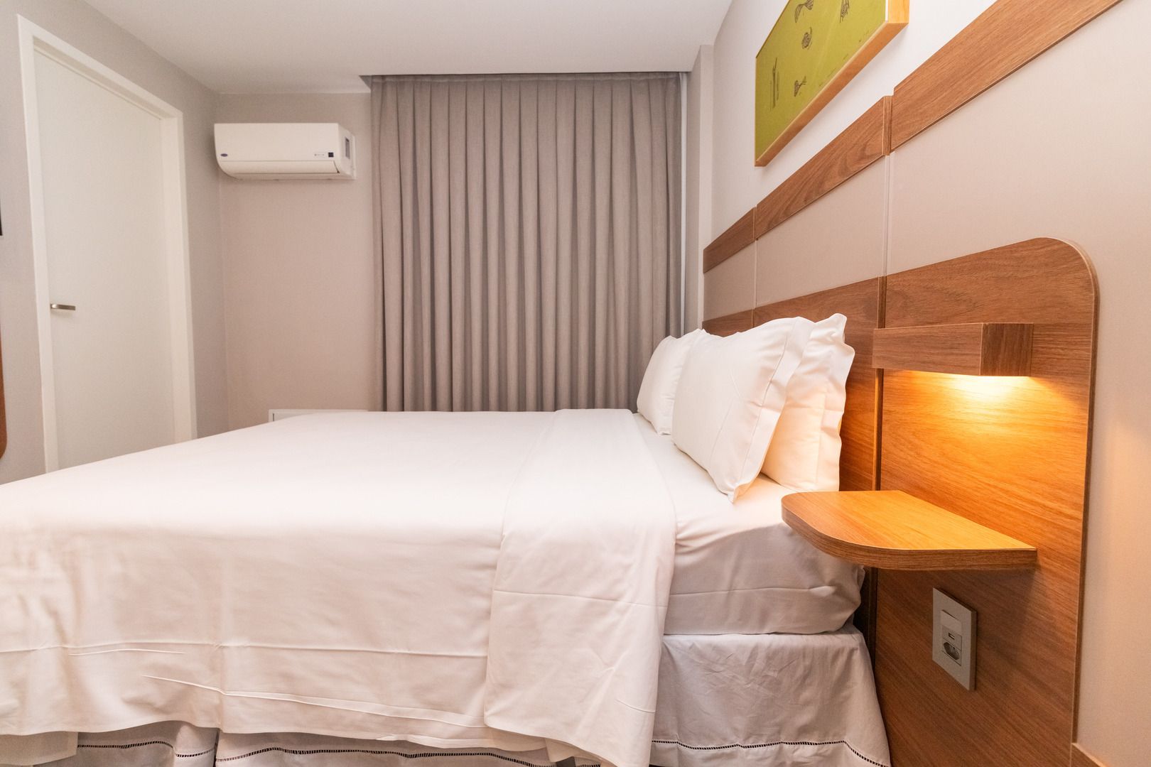 Quarto aconchegante com cama branca, cabeceira de madeira, abajur, cortinas e ar-condicionado.