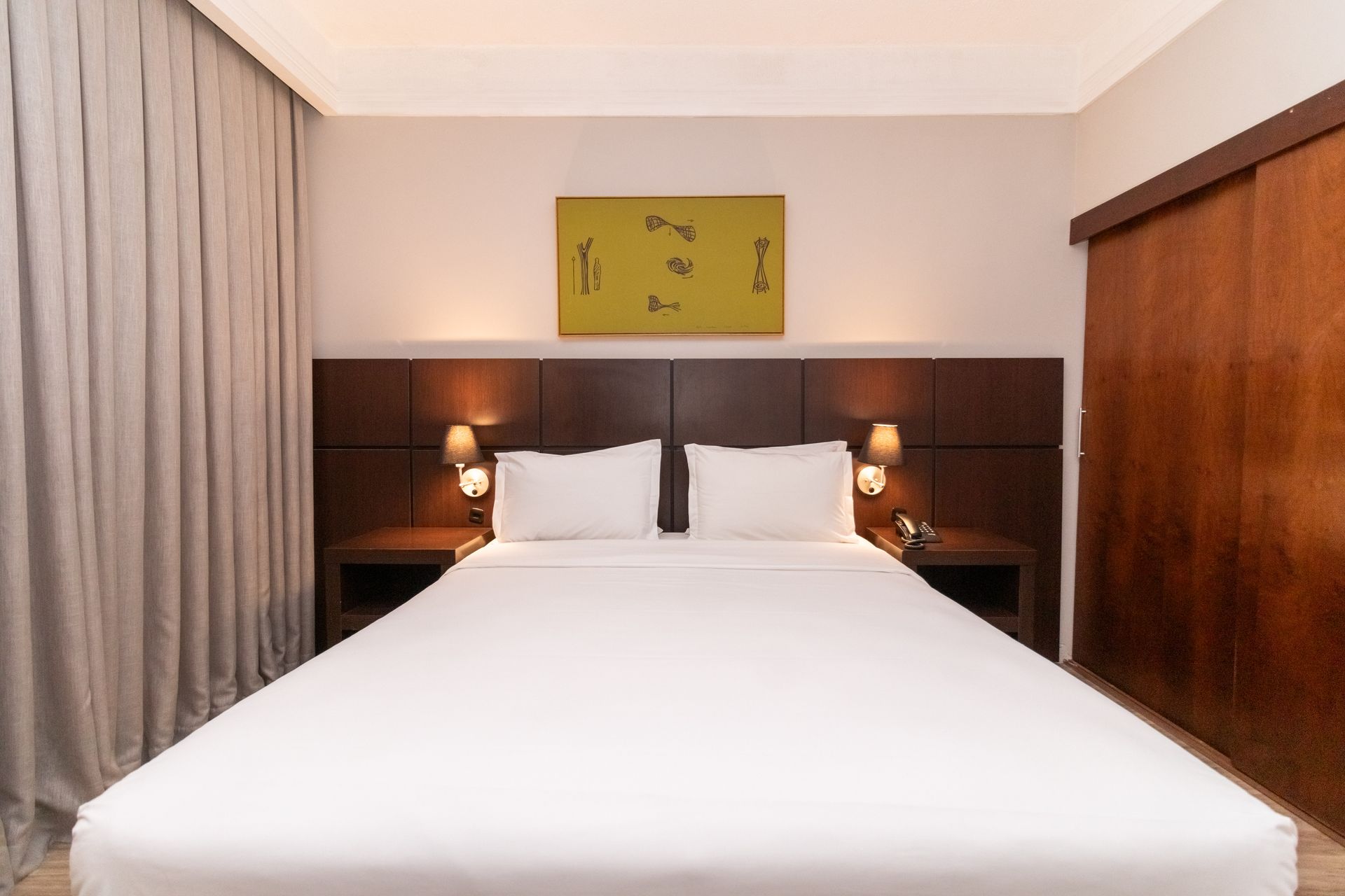 Quarto de hotel moderno com cama arrumada, cabeceira de madeira, candeeiros de cabeceira e cortinas.