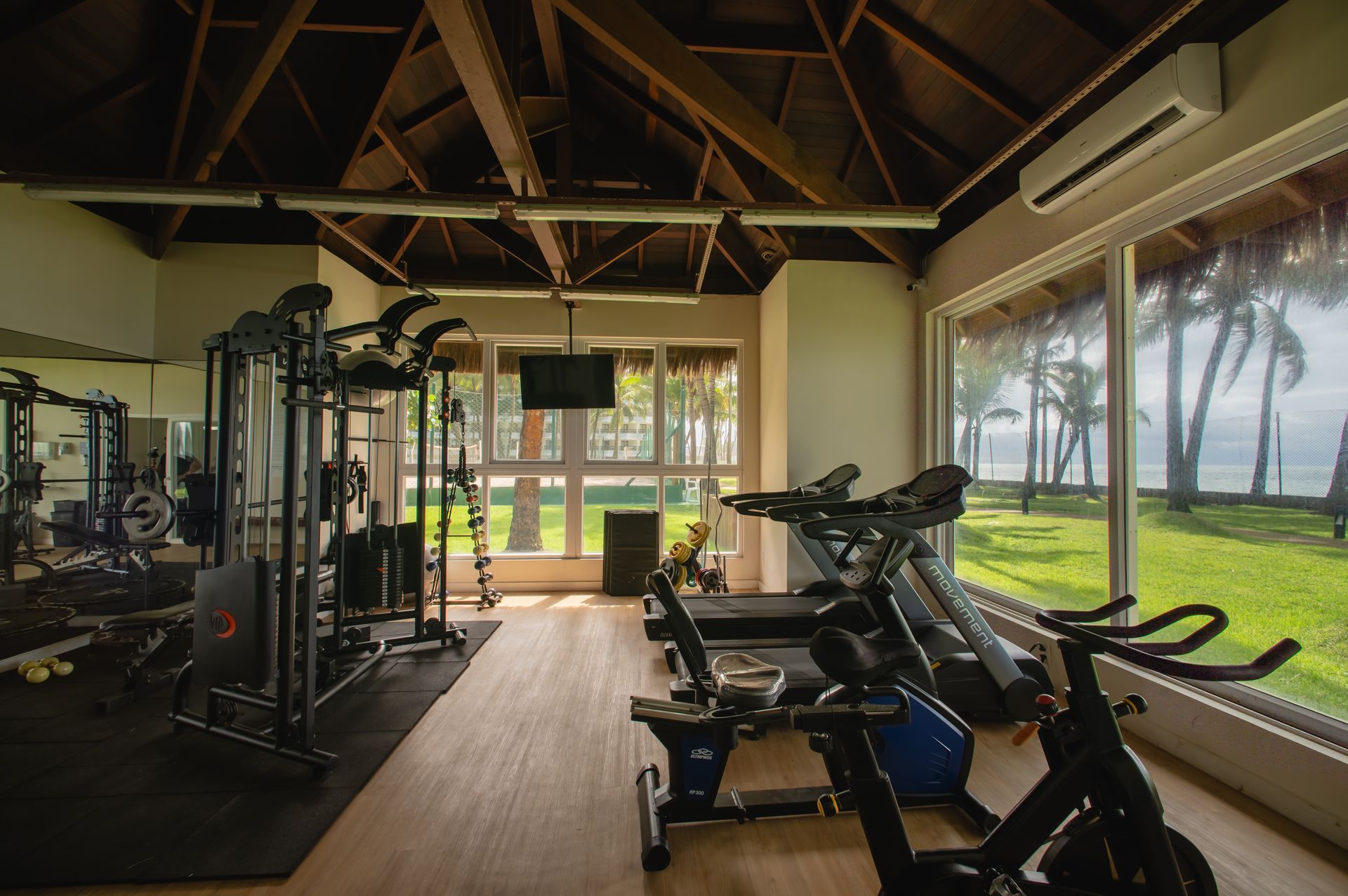Academia moderna com esteiras, aparelhos de musculação e grandes janelas com vista para uma área externa gramada.