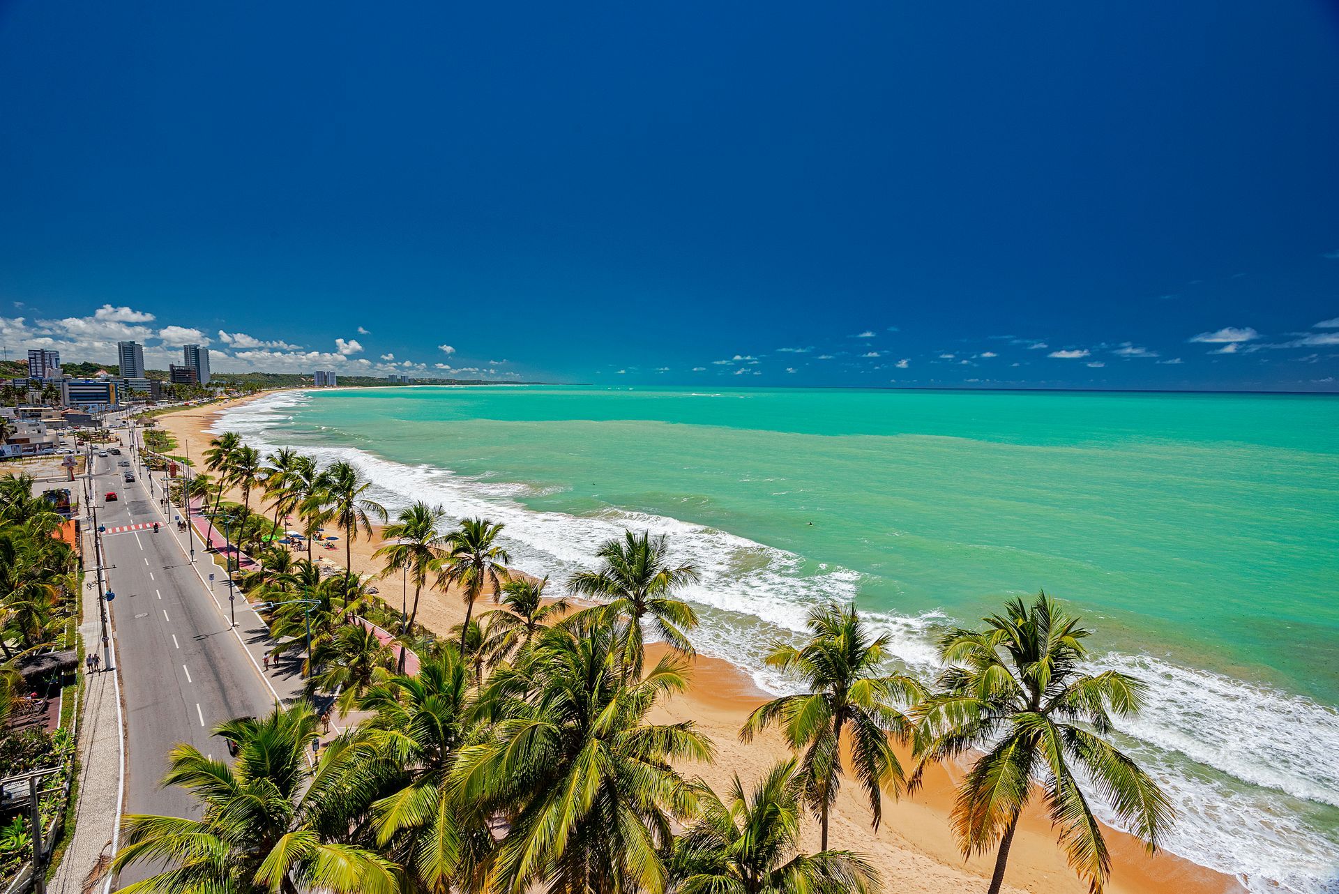 Vista aérea de uma praia ladeada por palmeiras, com mar turquesa e estrada costeira sob um céu azul límpido.