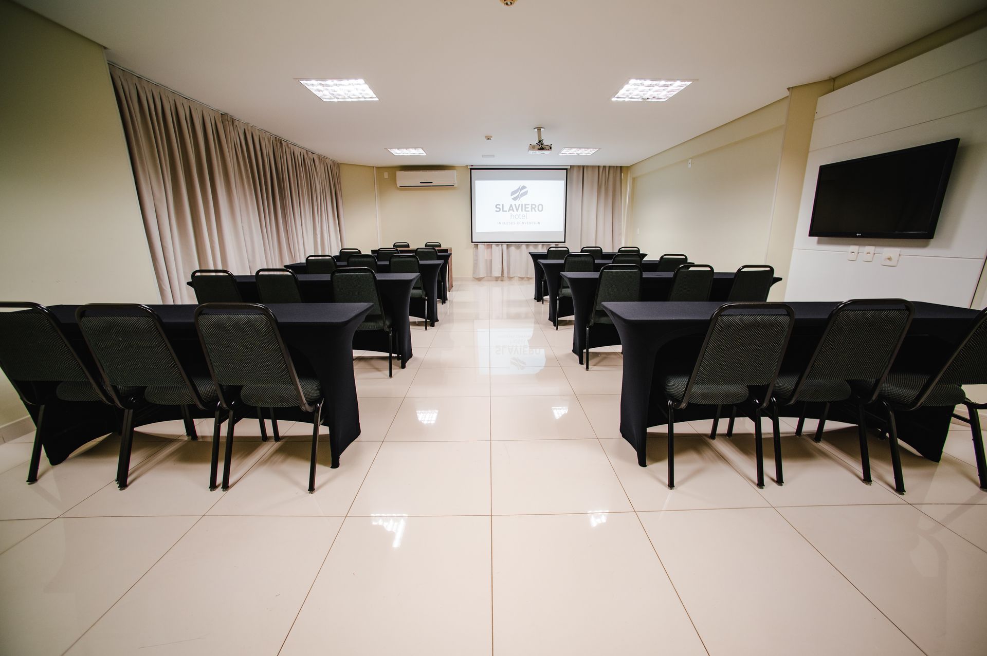 Sala de conferências vazia com fileiras de cadeiras pretas voltadas para uma tela de projeção.