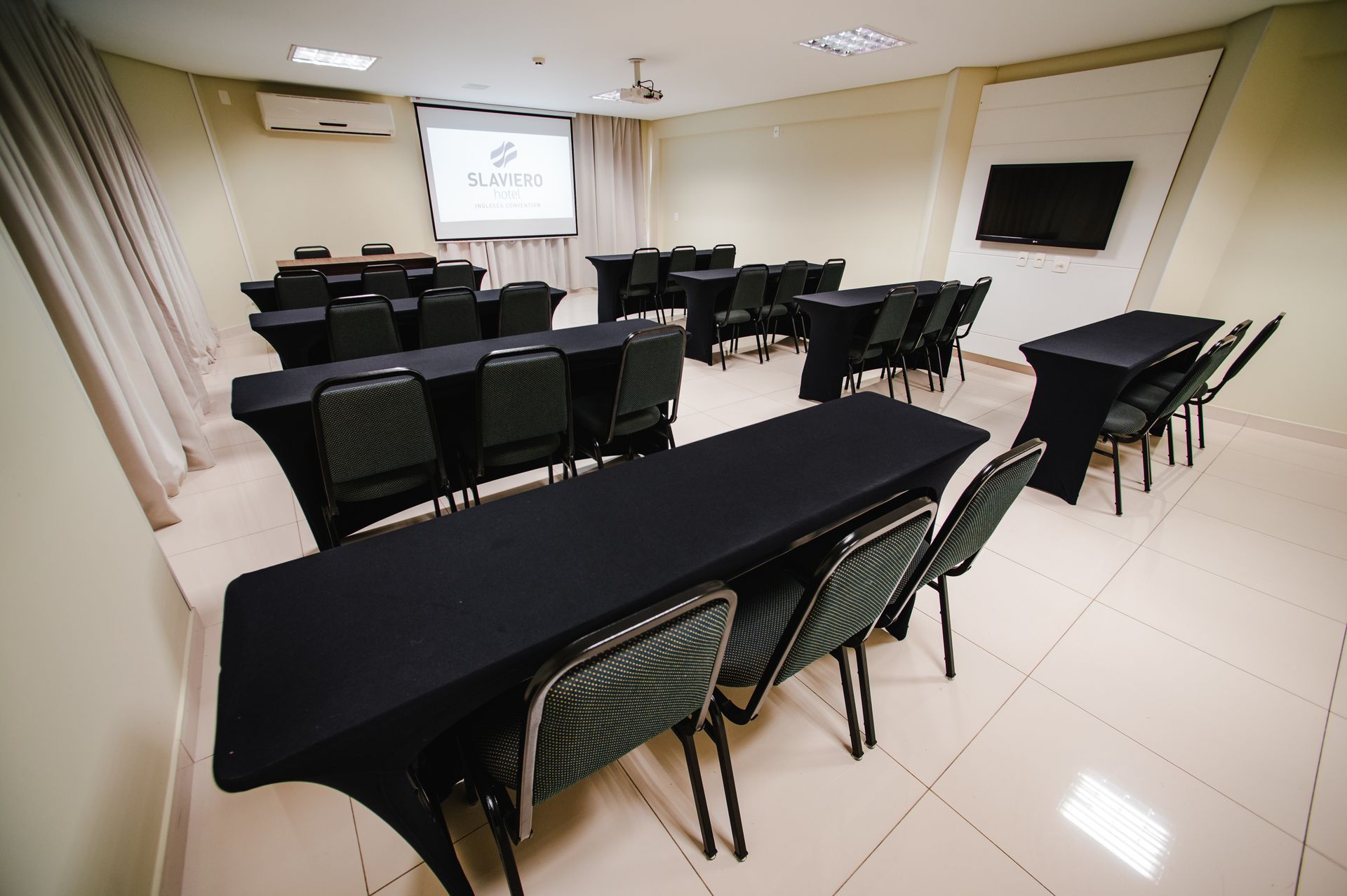 Sala de conferências com mesas e cadeiras revestidas de preto, tela de projeção e TV fixada na parede.