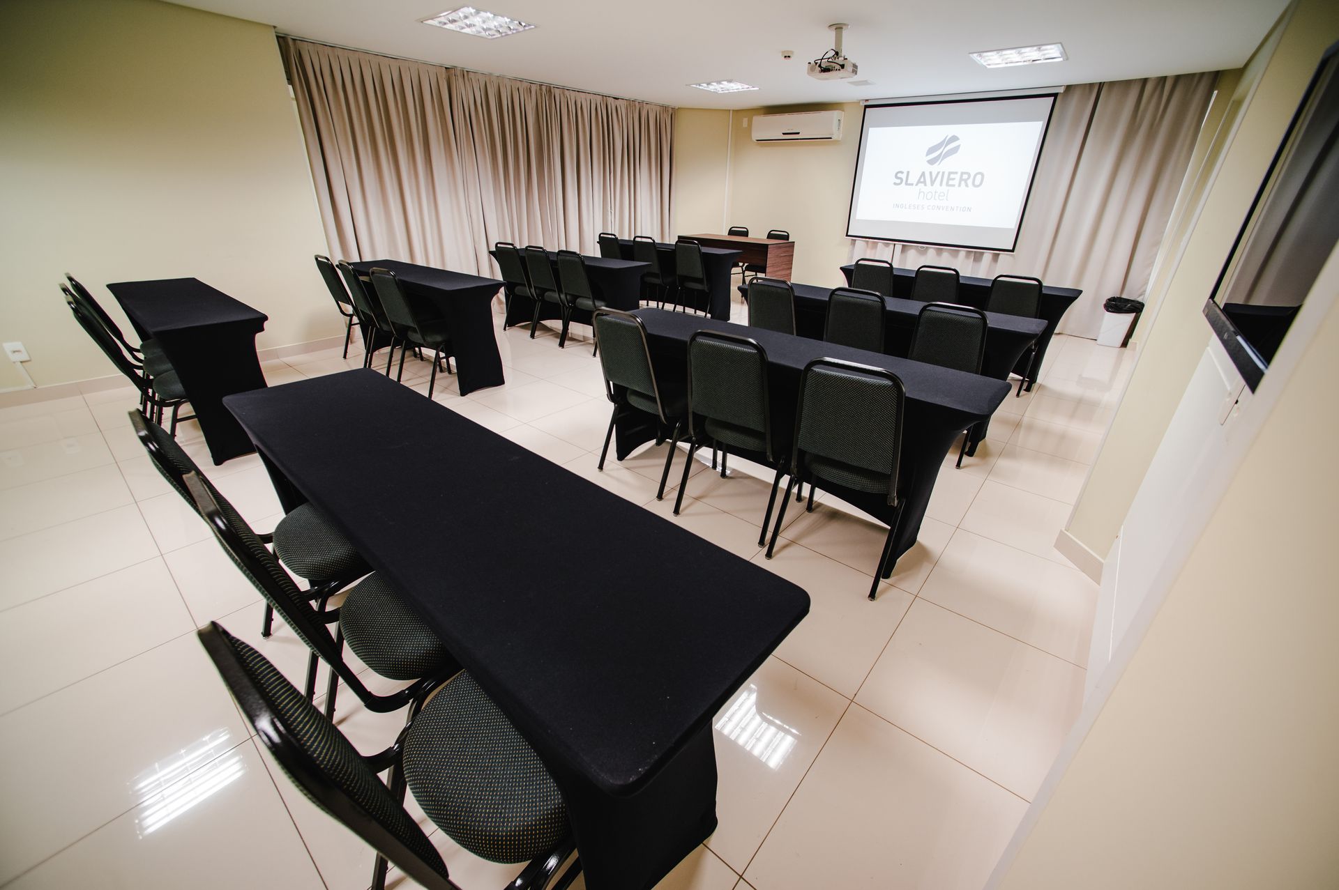 Sala de conferências vazia com mesas e cadeiras pretas, paredes brancas e uma tela de projeção na frente.