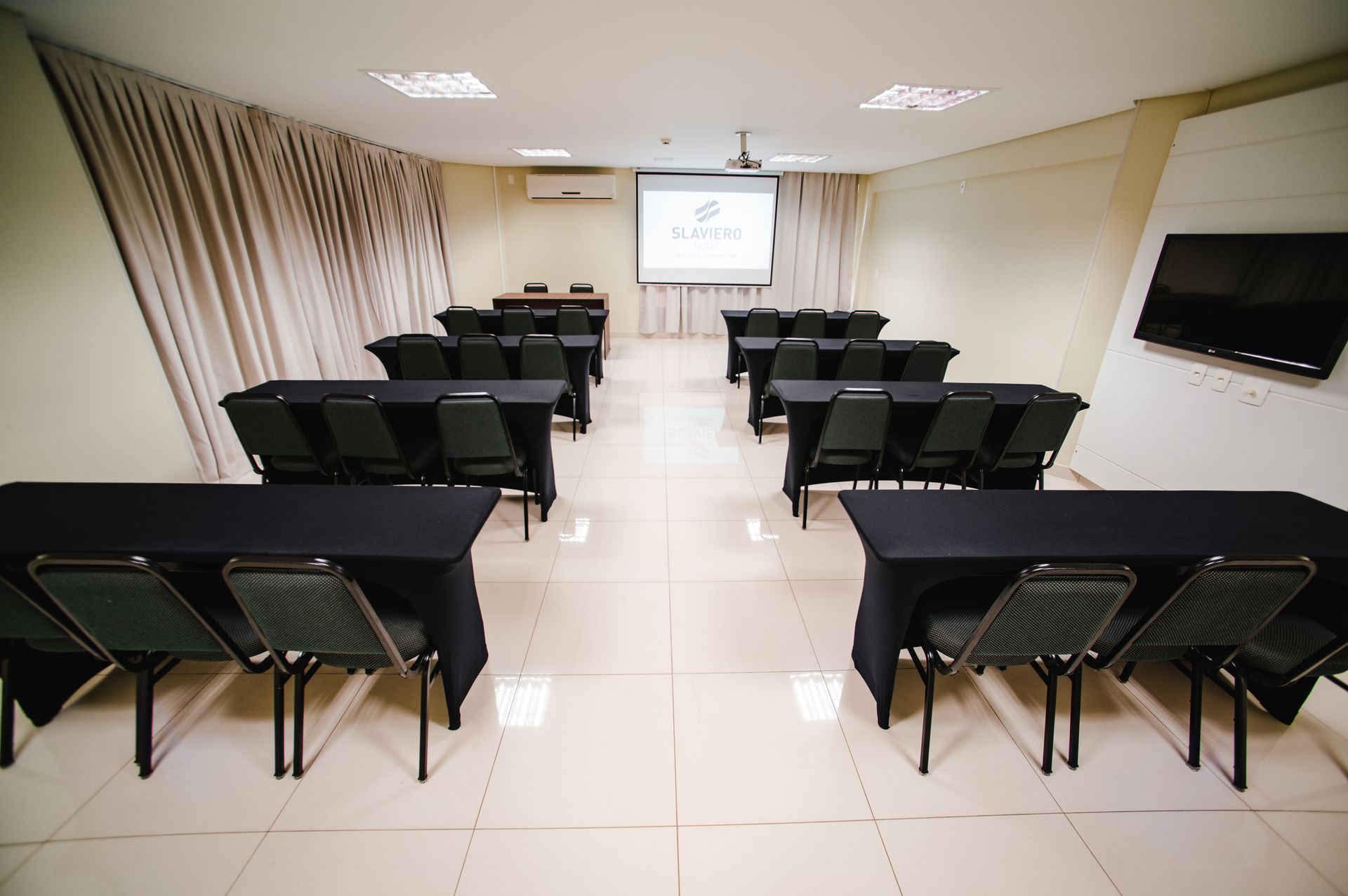 Sala de reuniões vazia com fileiras de cadeiras pretas voltadas para uma tela de projeção na frente.