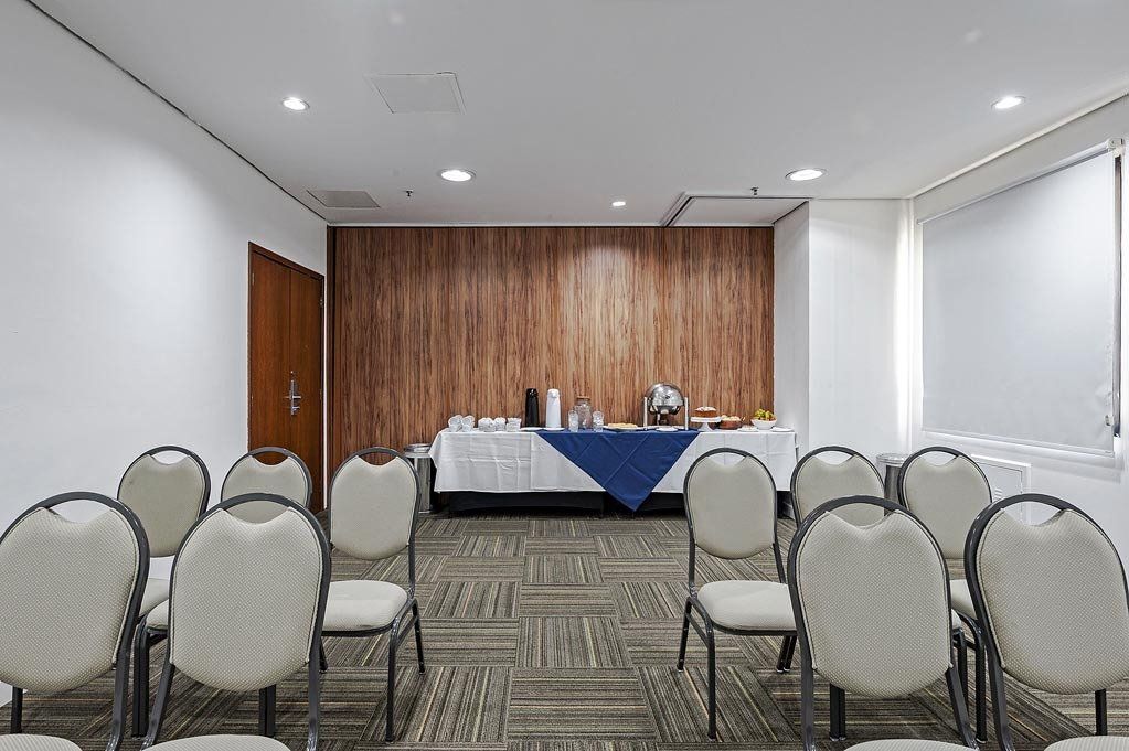 Sala de conferências vazia com fileiras de cadeiras e uma mesa coberta com uma toalha azul na frente.