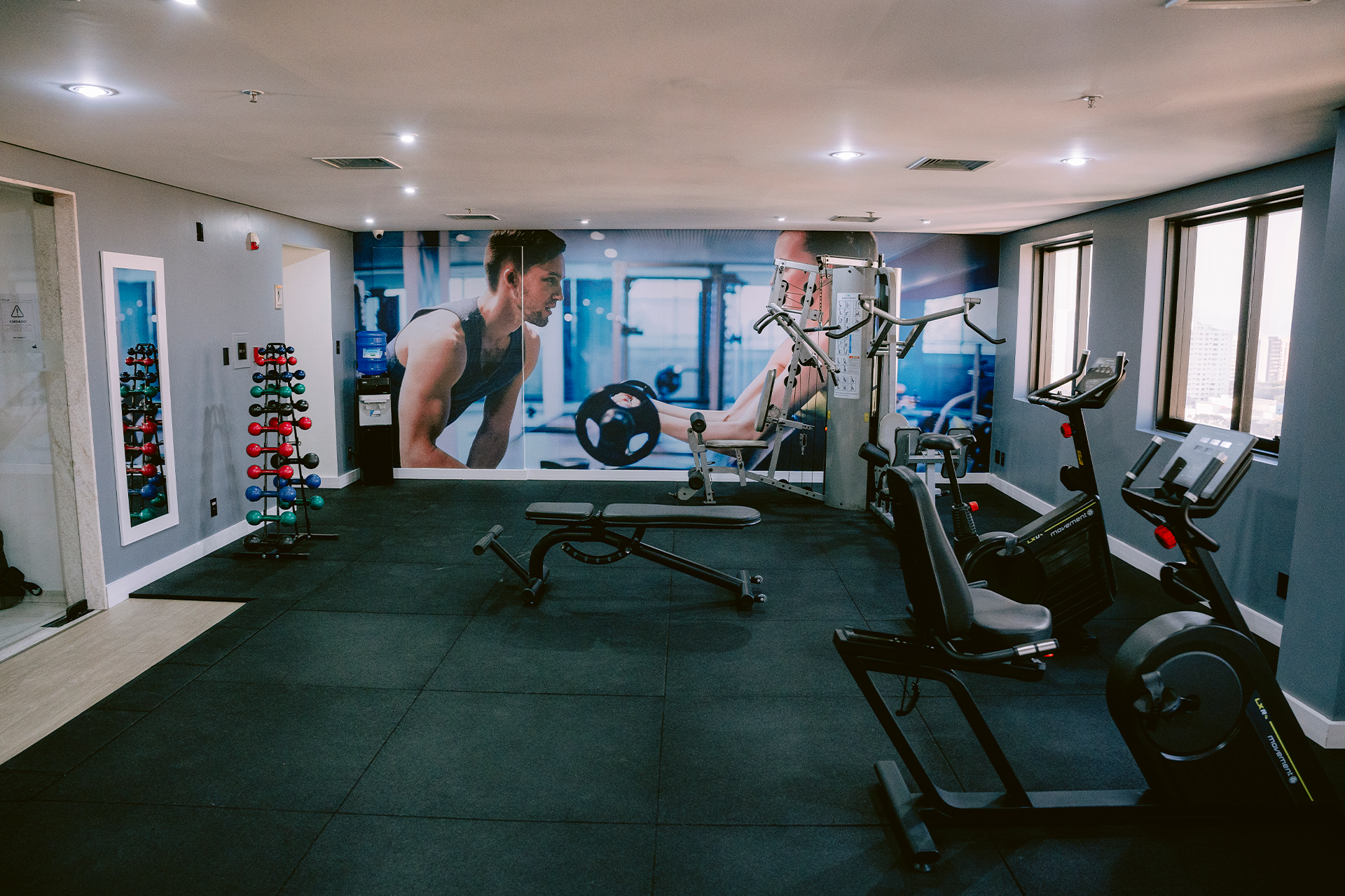 Interior de academia moderna com mural na parede, banco de exercícios, pesos e bicicletas ergométricas.