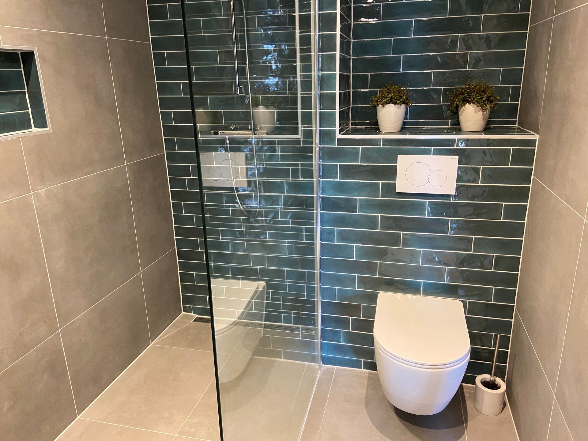 Moderne badkamer met donkergroene tegels, glazen douchecabine, wit toilet en planken met planten. 
