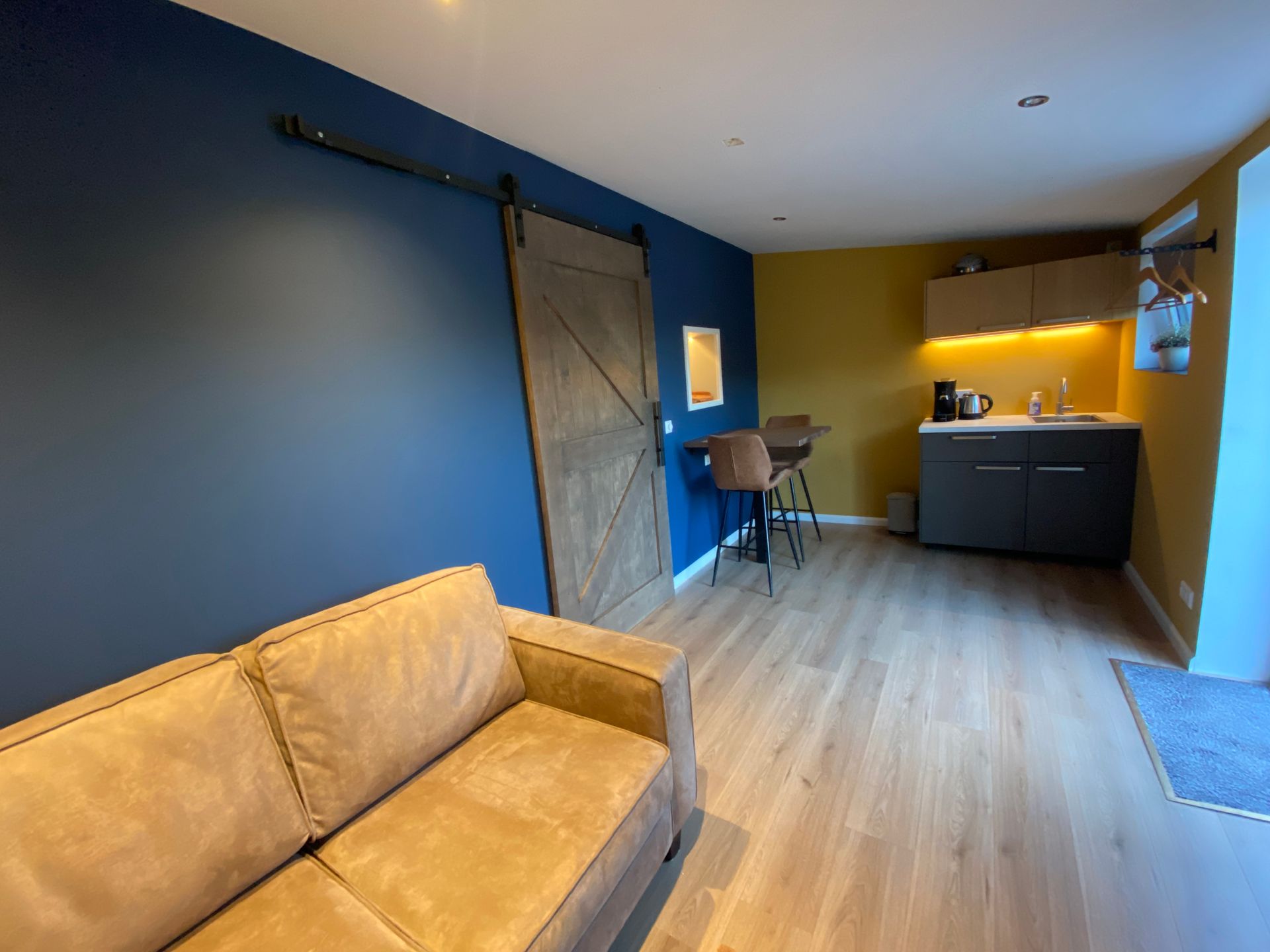 Moderne woonkamer met blauwe accentmuur, beige bank, schuifdeur in boerderijstijl en een kleine keukenhoek.