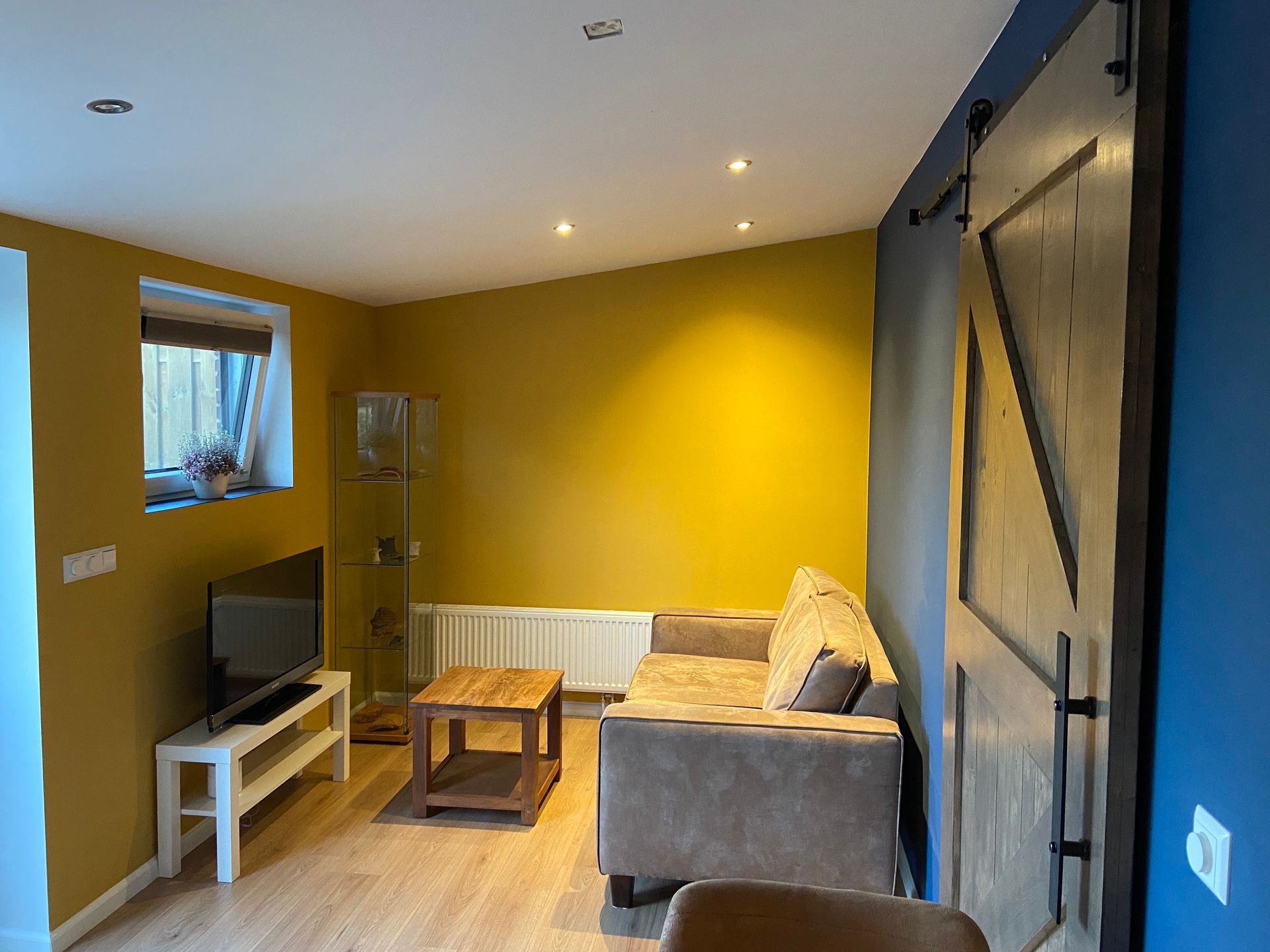 Kleine woonkamer met gele muren, grijze bank, houten salontafel, tv-meubel en schuifdeur in landelijke stijl.