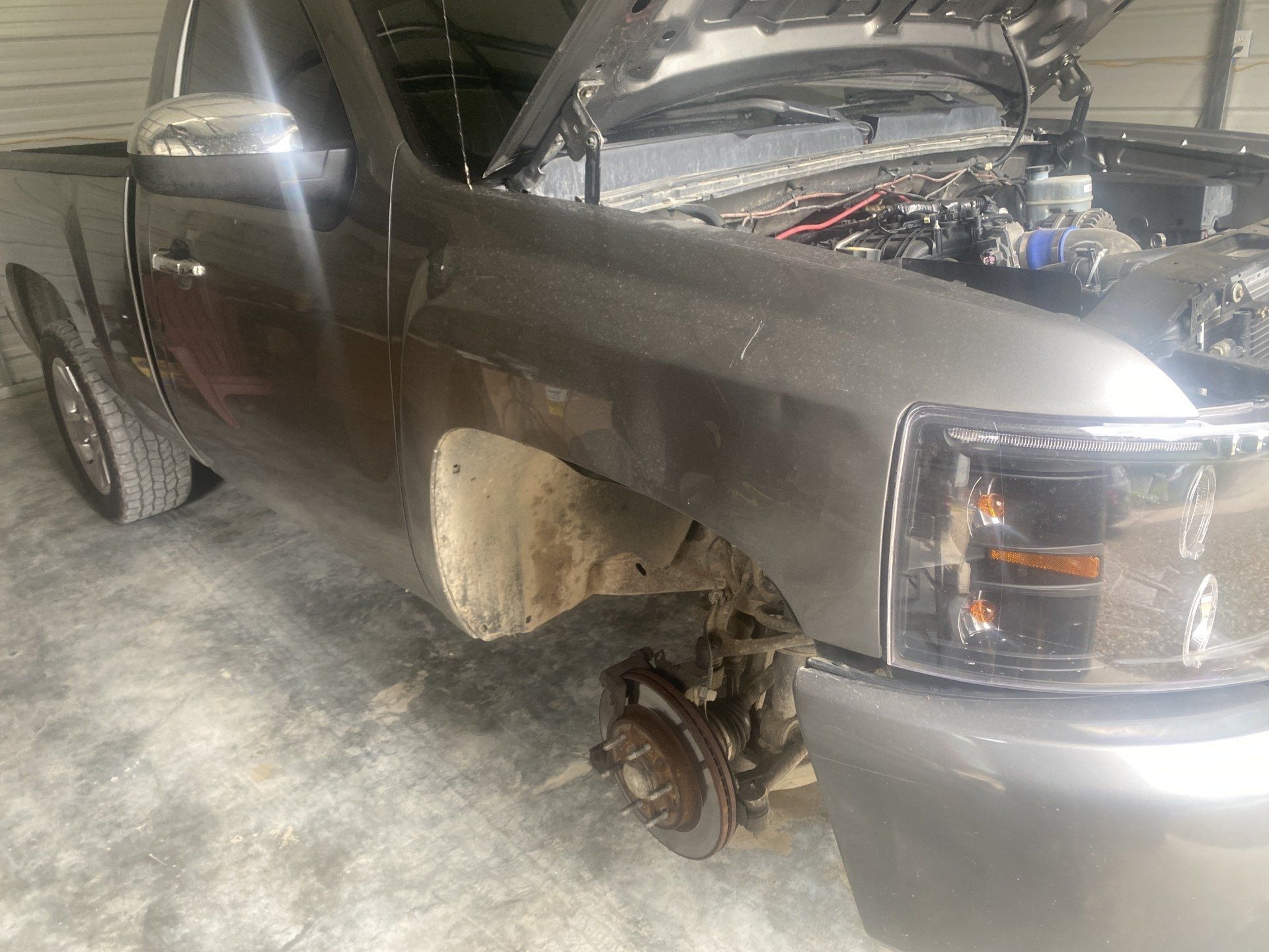 A gray truck with a broken wheel is parked in a garage with the hood open.
