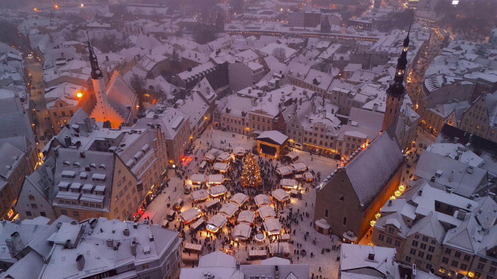 Snow-covered town square with Christmas market, illuminated stalls, and central tree. Buildings surround the plaza.