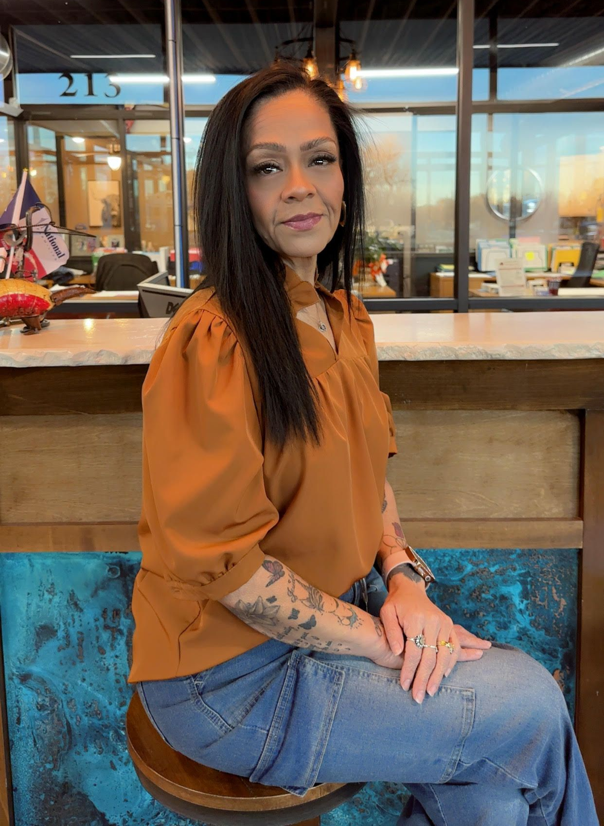 Woman in an orange blouse and jeans seated indoors at a café counter, with a windowed background.