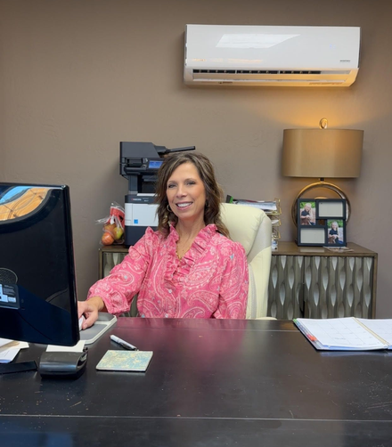 Woman seated at an office desk in a pink blouse, smiling beside a computer and papers.