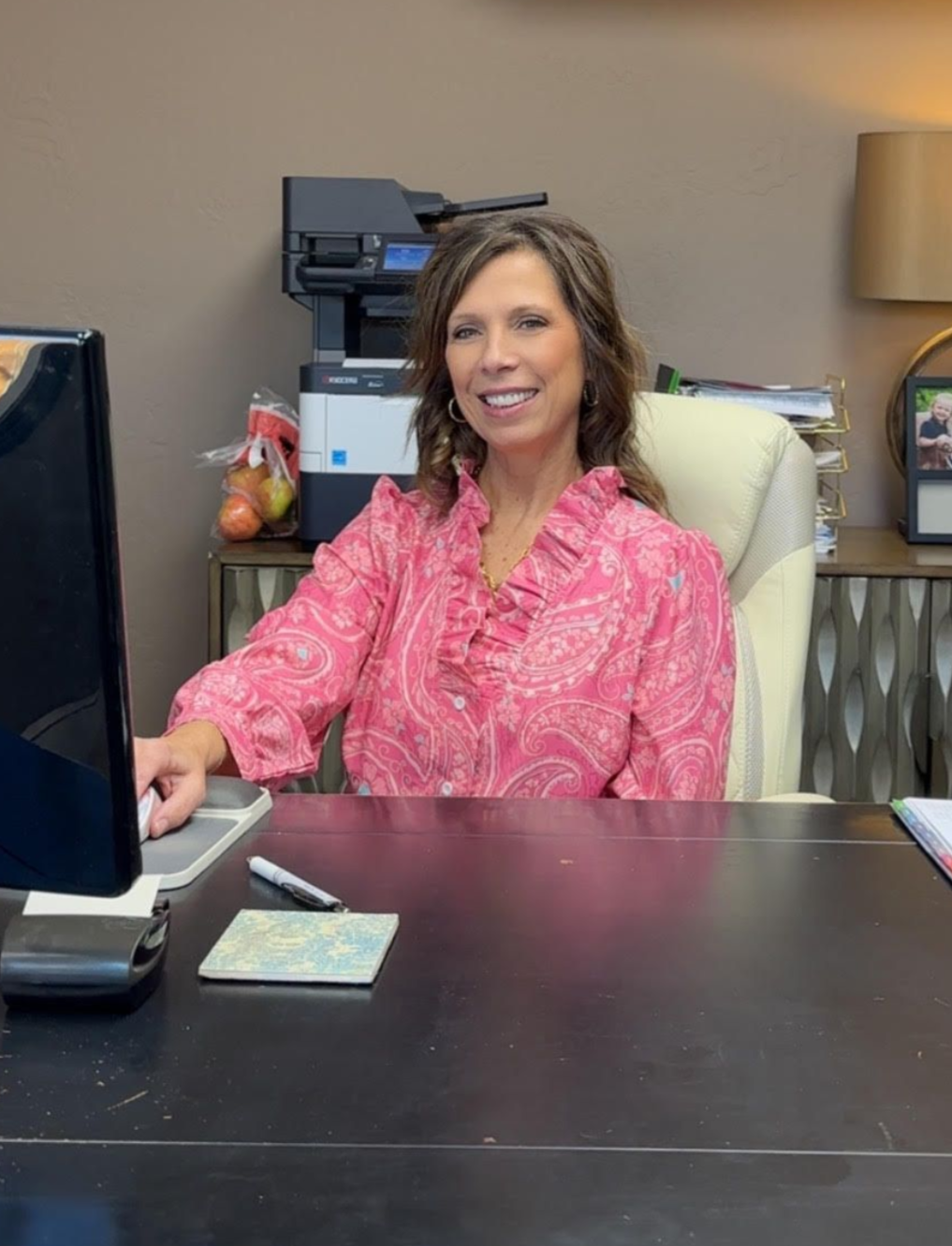 Woman in a pink blouse smiling at an office desk with a computer and papers.