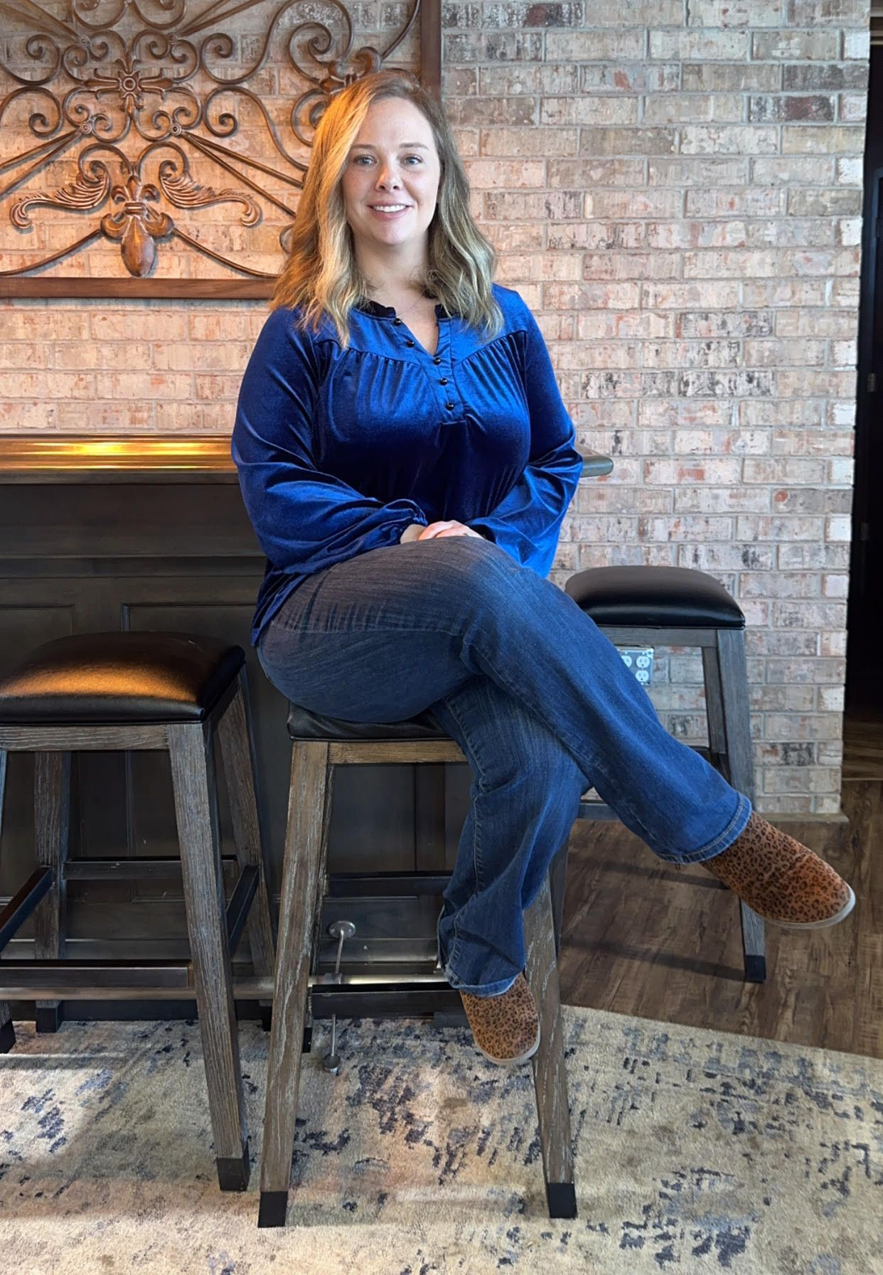 Woman in a blue blouse and jeans sitting on a barstool in front of a brick wall and fireplace.