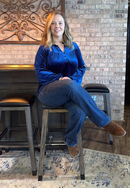 Woman in a blue blouse sitting on a high stool in a brick-walled room by a fireplace