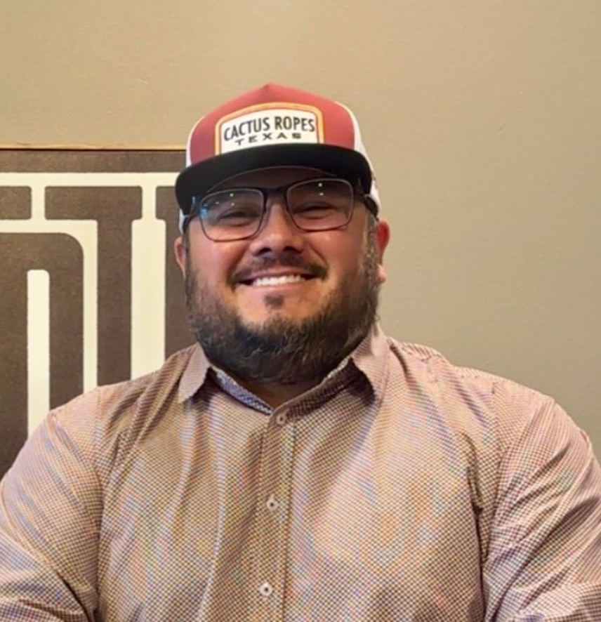 Smiling man in glasses and a Cactus Ropes cap, wearing a patterned button-up shirt indoors