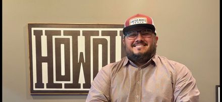Man smiling in a cap and glasses standing beside a “HOME” sign on a beige wall.