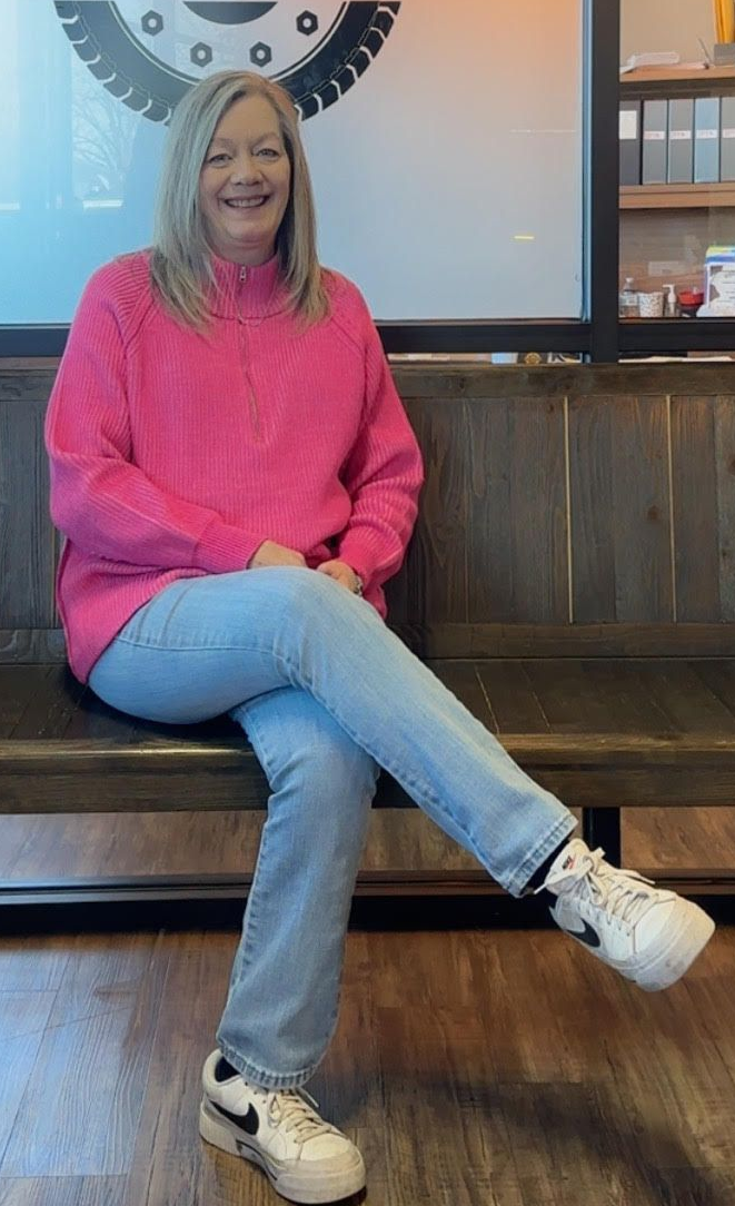 Woman in a bright pink sweater and jeans sitting cross-legged on a wooden bench indoors.