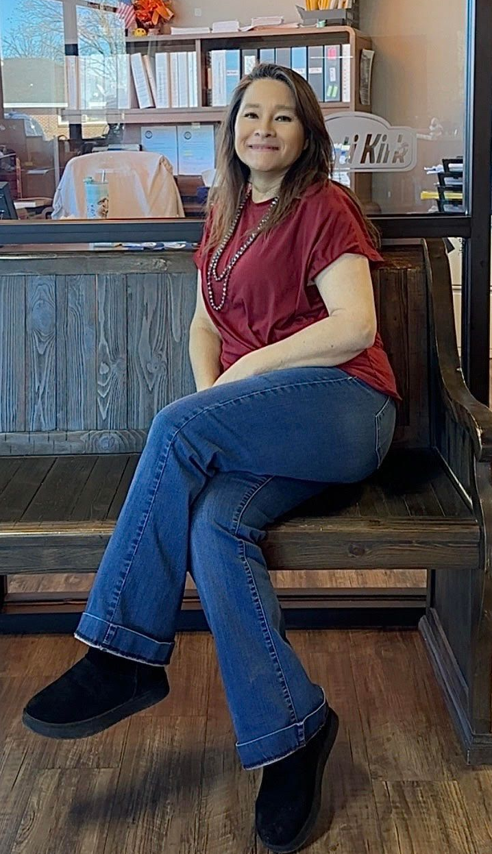 Woman in a red shirt and jeans sitting on a wooden bench in a café, smiling at the camera.