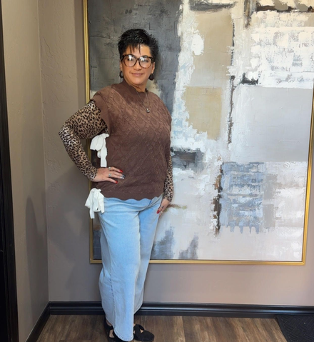 Person posing indoors beside a large abstract painting, wearing a brown sweater and light blue jeans.