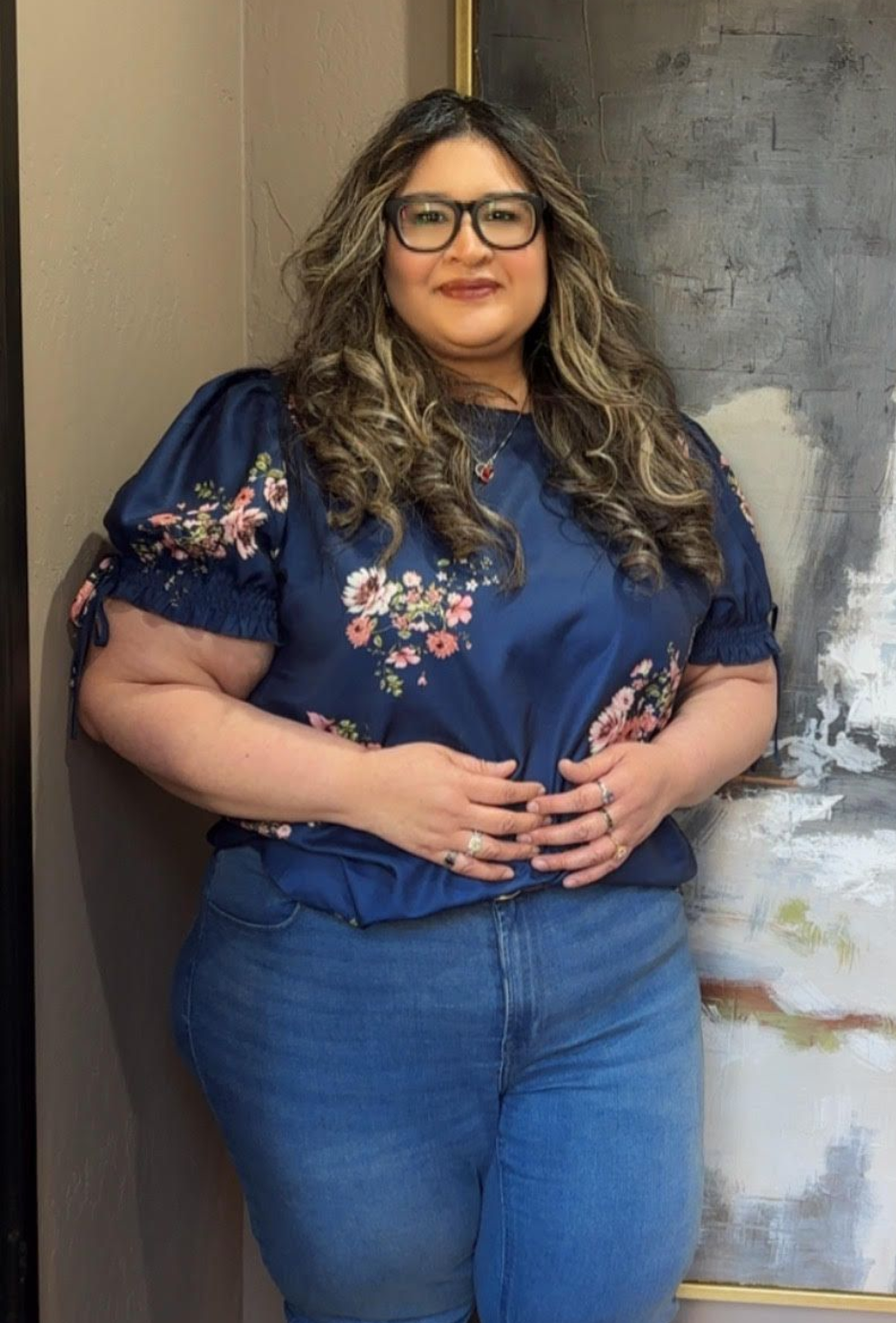 Woman standing indoors, wearing a navy floral blouse and jeans, hands clasped, near a weathered doorway.