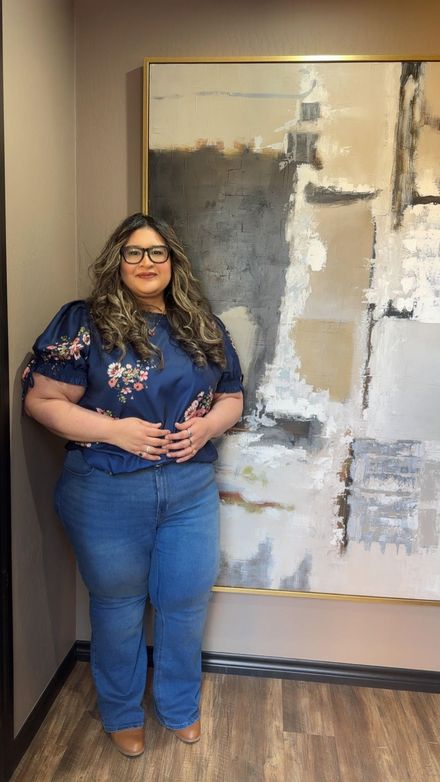 Woman in glasses stands indoors beside a large abstract painting, wearing a navy floral top and blue jeans.