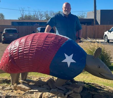 Man standing beside a large red, blue, and gray armadillo sculpture in a yard