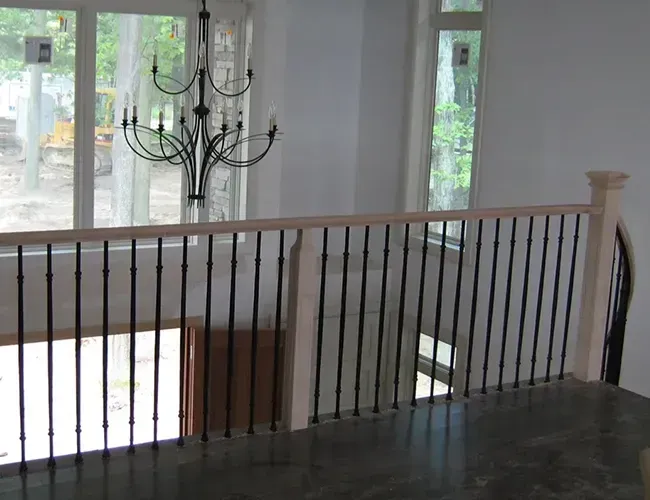 Indoor staircase landing with black metal railing and chandelier, overlooking large windows and greenery outside