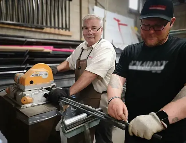 Two workers operating a metal-cutting saw in a workshop, one in gloves, with stacked materials behind them