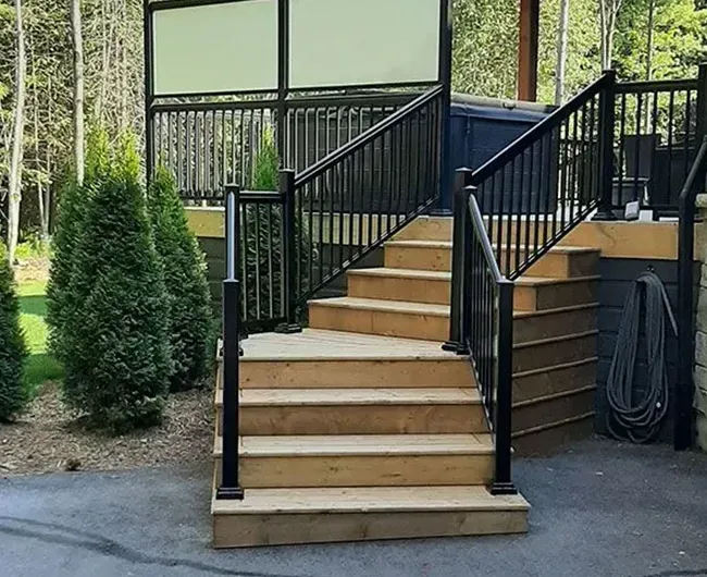 Wooden outdoor stairs with black railings leading to a deck beside a house, next to two evergreen shrubs.