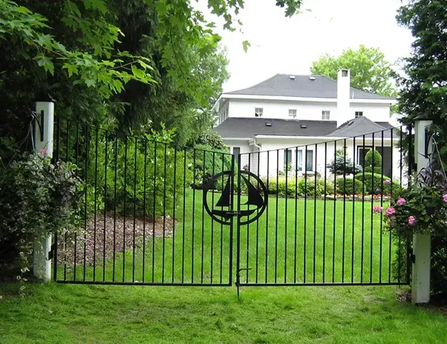 Black metal gate with circular emblem in a green garden, leading to a white house