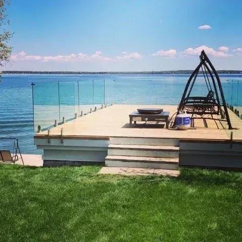 Lakeside deck with glass railing, steps, lawn, lounge chairs, and an arched fire pit by calm blue water.