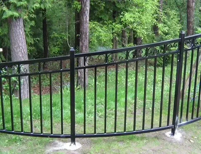 Black metal fence curving along a grassy path beside trees in a wooded area