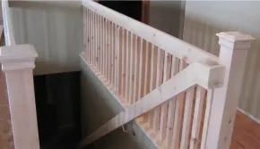 Unfinished light-colored wooden railing and stairwell with vertical balusters and two square posts.