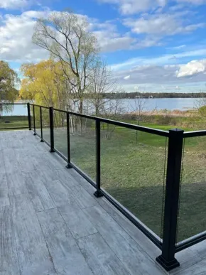 A deck with a black metal railing and glass panels overlooking a lake and trees under a blue sky with clouds.