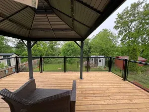 A wooden deck featuring a dark gazebo over a wicker lounge chair, overlooking a grassy yard with small sheds and trees.