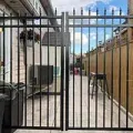 Black metal gate blocking a narrow backyard walkway between a house and wooden fence