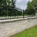 Black wrought-iron fence atop a light stone wall beside a green lawn and trees