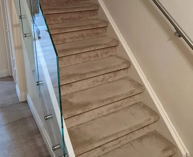 Beige carpeted staircase with glass railing and white walls in a hallway