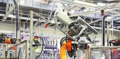 An automotive factory robot assembling a car frame. The robot is orange, and the frame is silver.