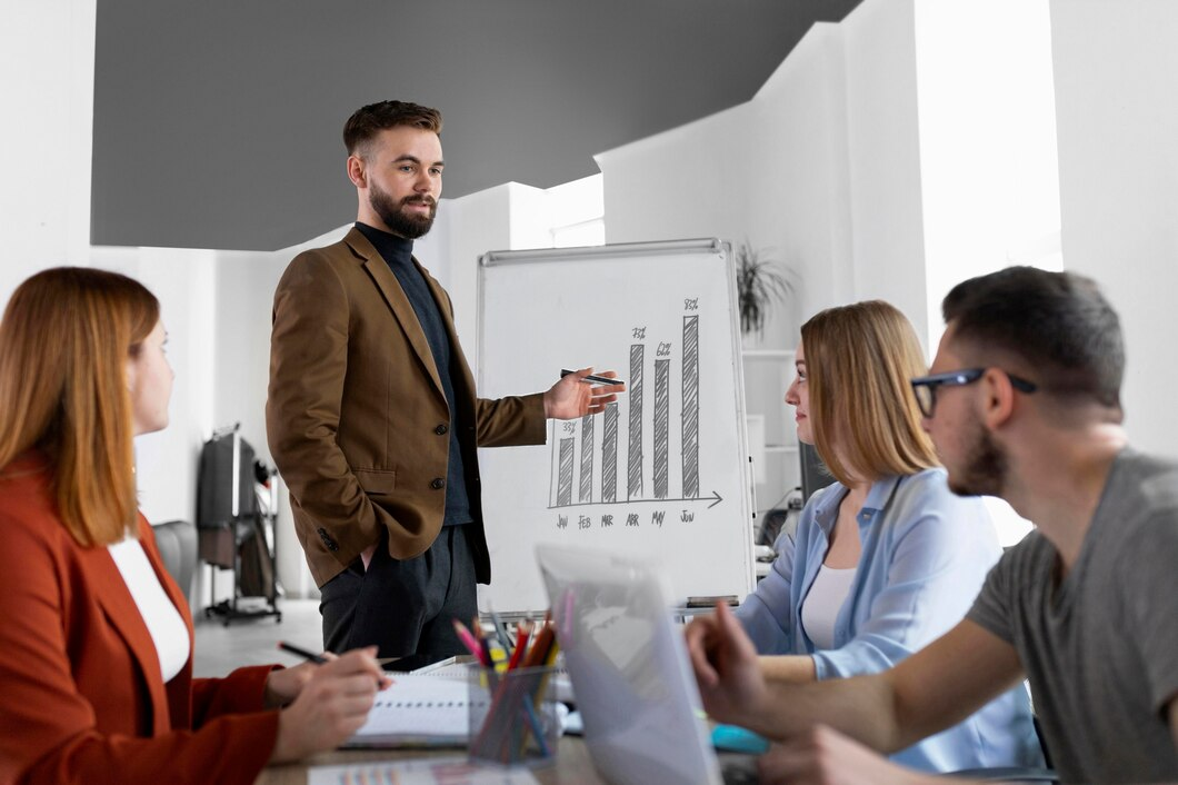 A man in a blazer presents data to a group in an office setting.