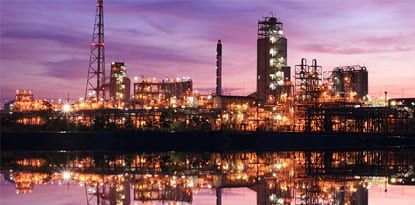 Illuminated industrial plant at dusk, reflected in water. Purple and orange hues dominate.
