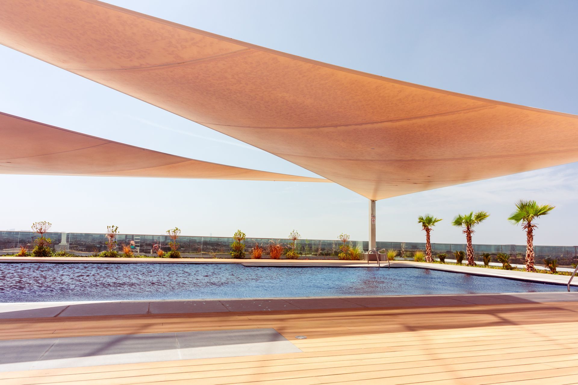 Modern outdoor pool with sun-shade sails and palm trees on a sunny deck.