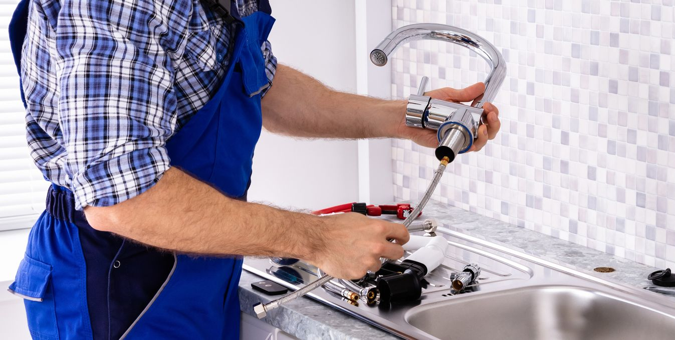 Plumber in blue overalls fixing a kitchen sink faucet with tools, against a tiled wall.