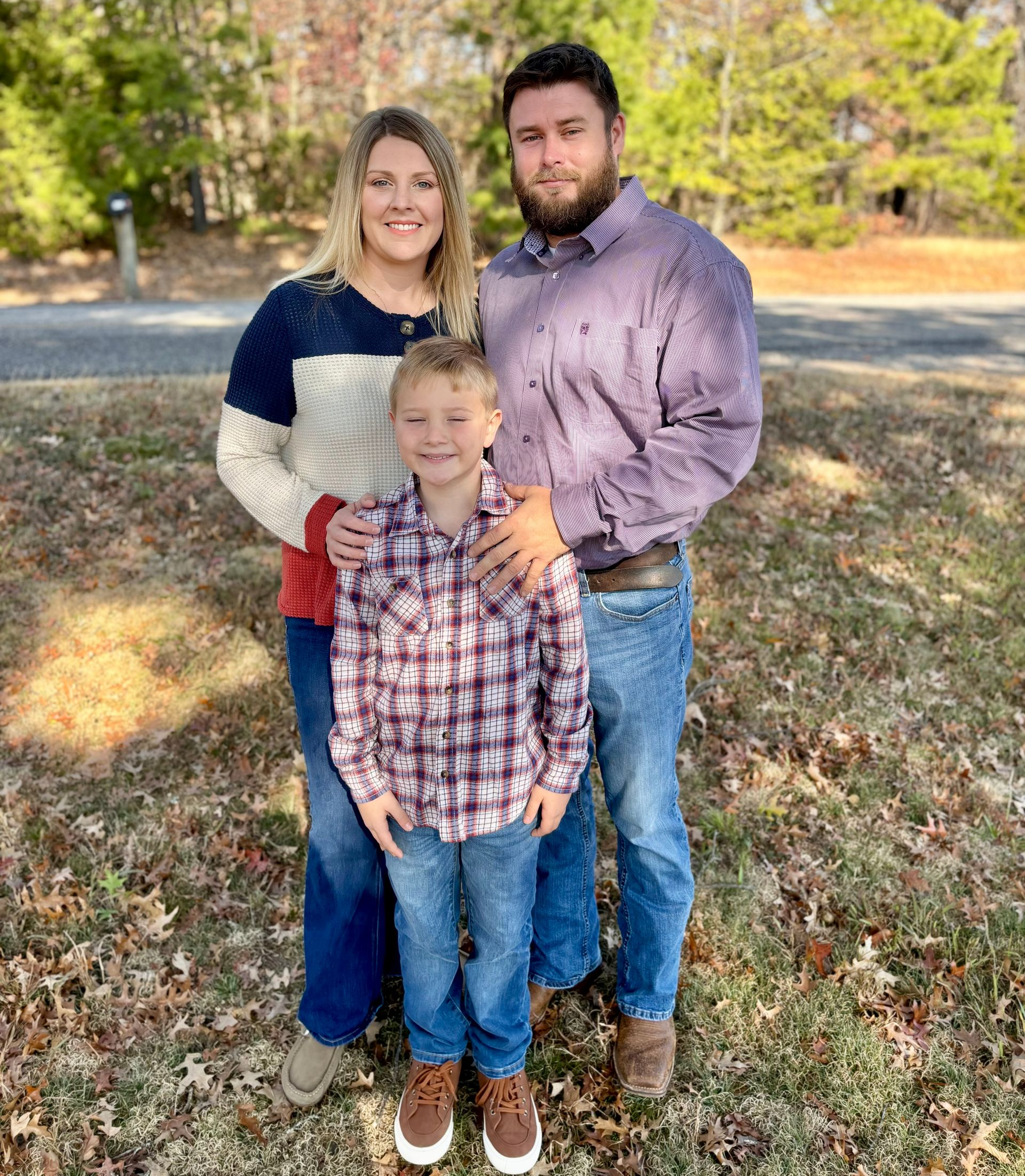 Family of three smiling outdoors: woman in sweater, man in button-down, boy in plaid, autumn setting.