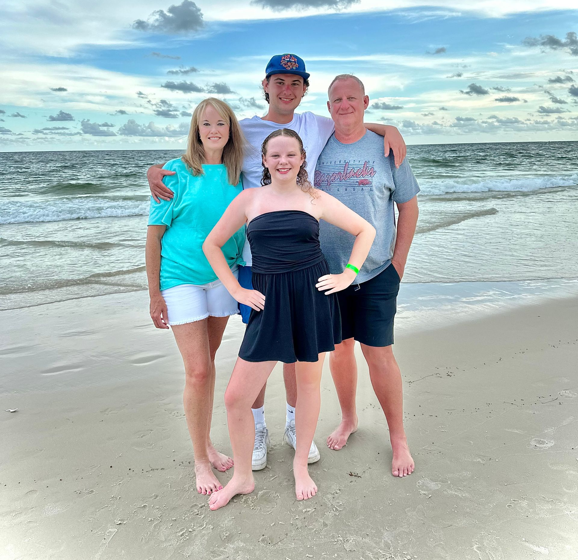 Family of four at the beach, smiling. Woman in turquoise top, man in blue hat, girl in black dress. Cloudy sky, ocean.