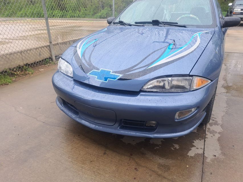 A blue car is parked in a parking lot next to a chain link fence.