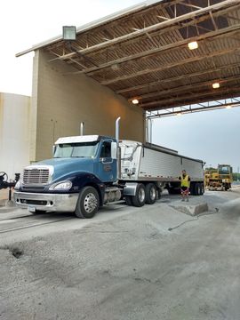 A blue semi-truck sits under an industrial canopy on a gravel lot as a person in a safety vest stands nearby.